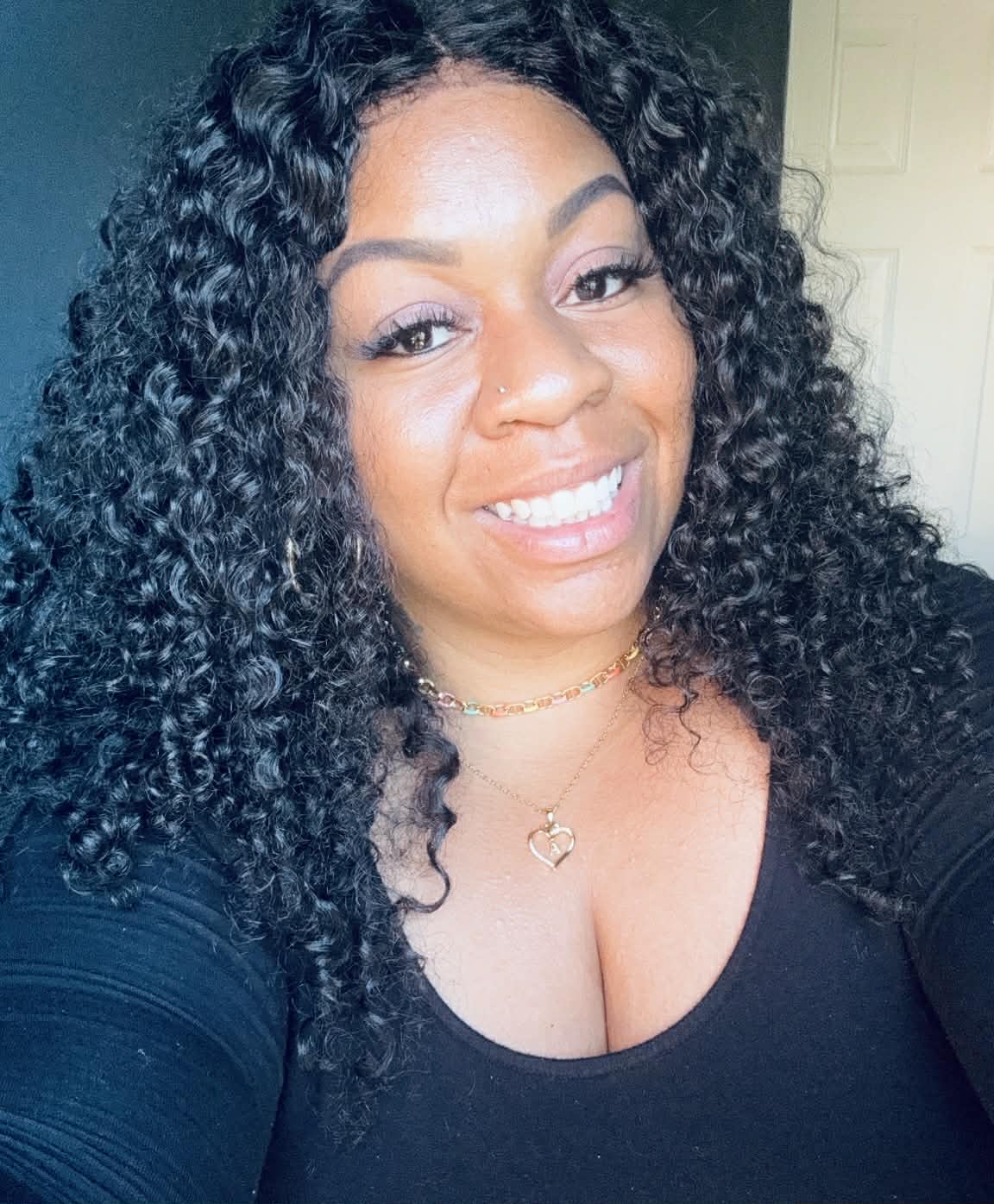 Smiling woman with voluminous curly black hair, wearing a black top and gold necklaces.