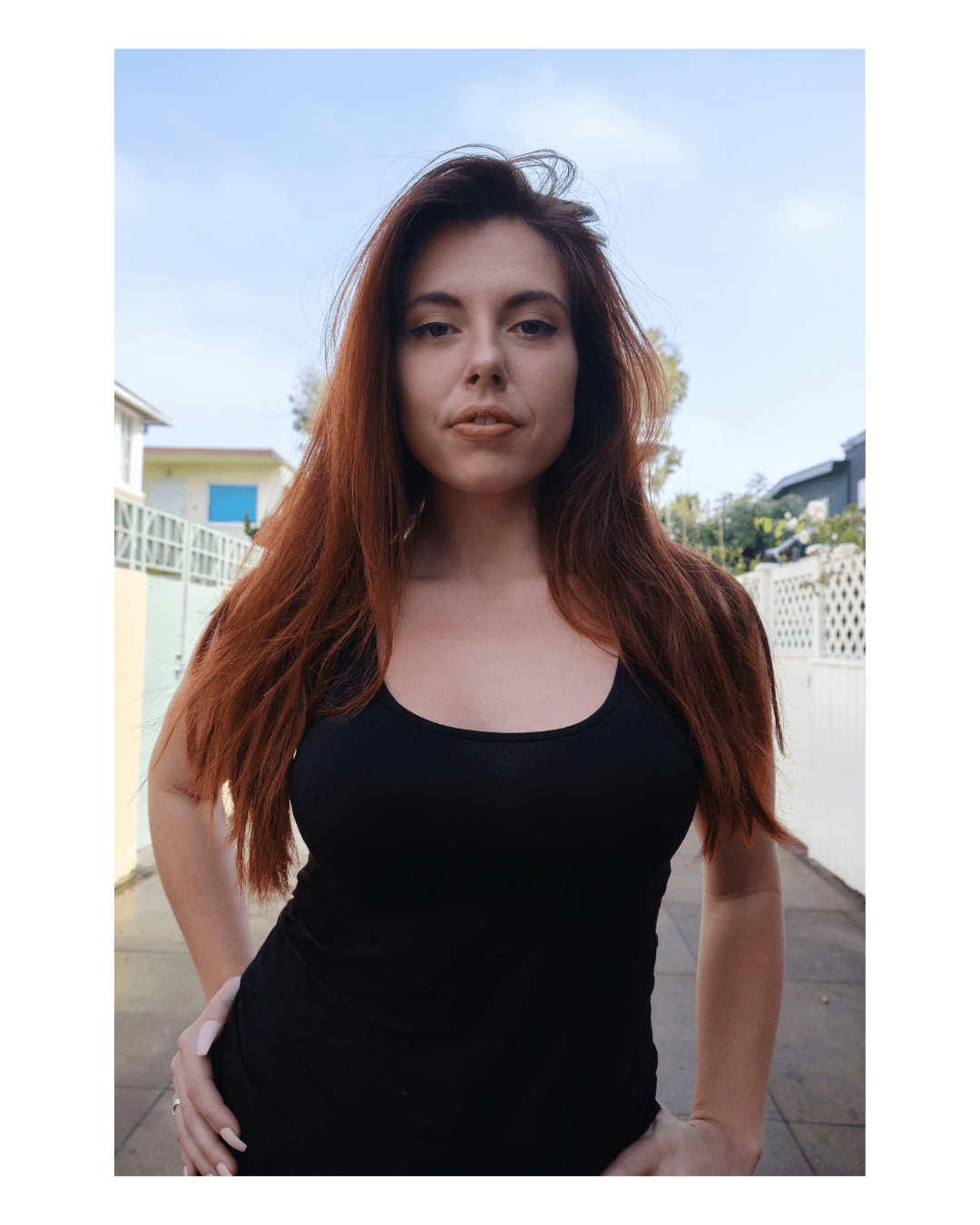 Redheaded woman in a black tank top posing outdoors with hands on her hips.