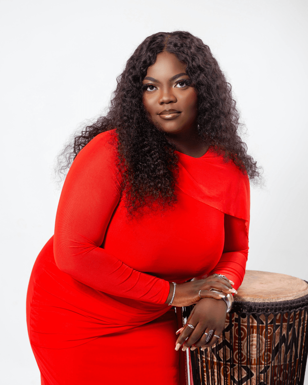 Black woman with long curly hair in a red dress leaning on an African drum.