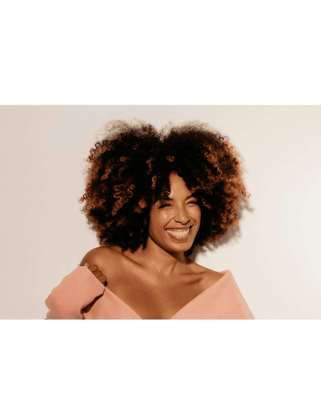 Smiling woman with voluminous curly hair and blonde highlights in a pink off-the-shoulder top.