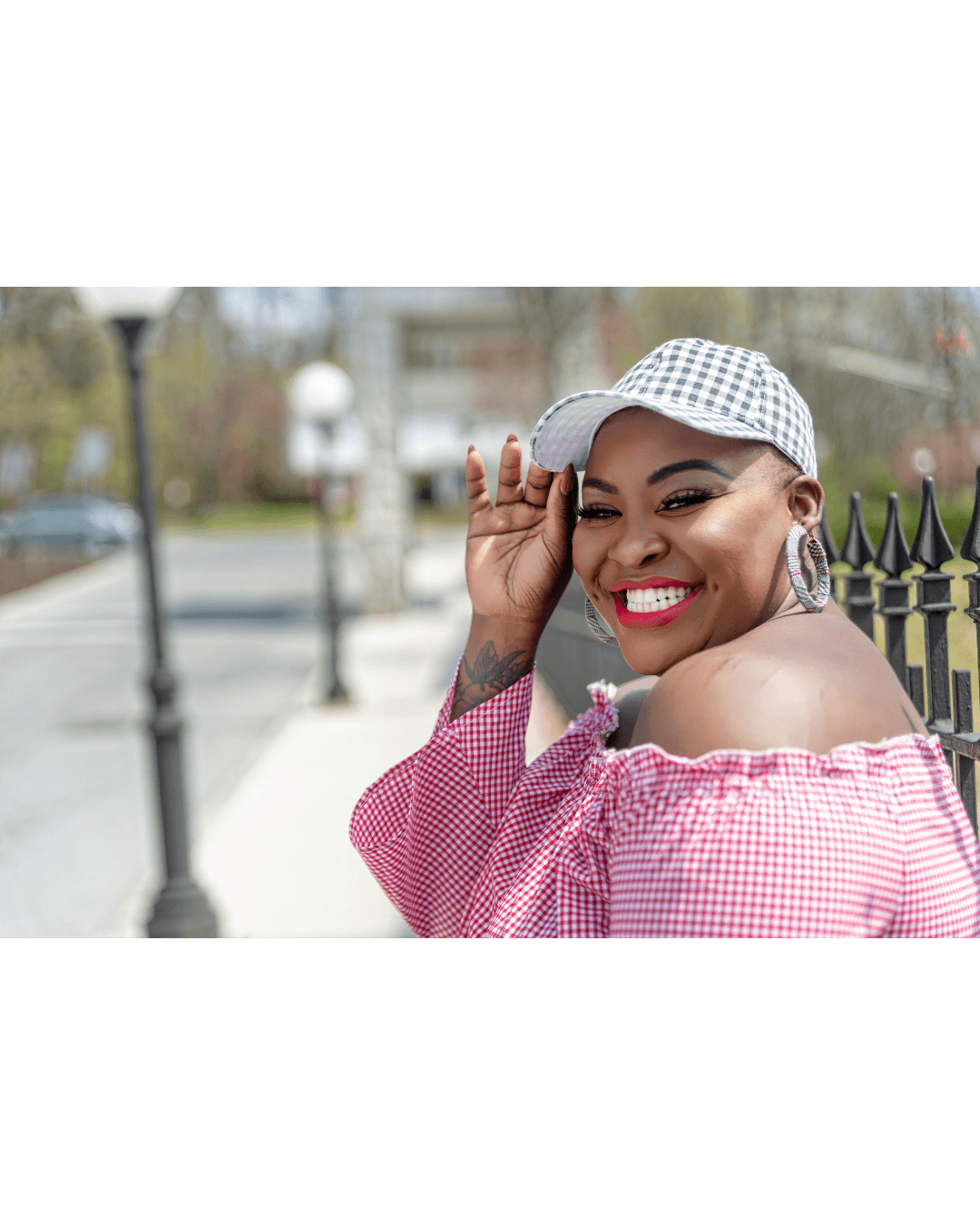 Smiling woman in red gingham top and checkered cap poses outdoors near a black fence.