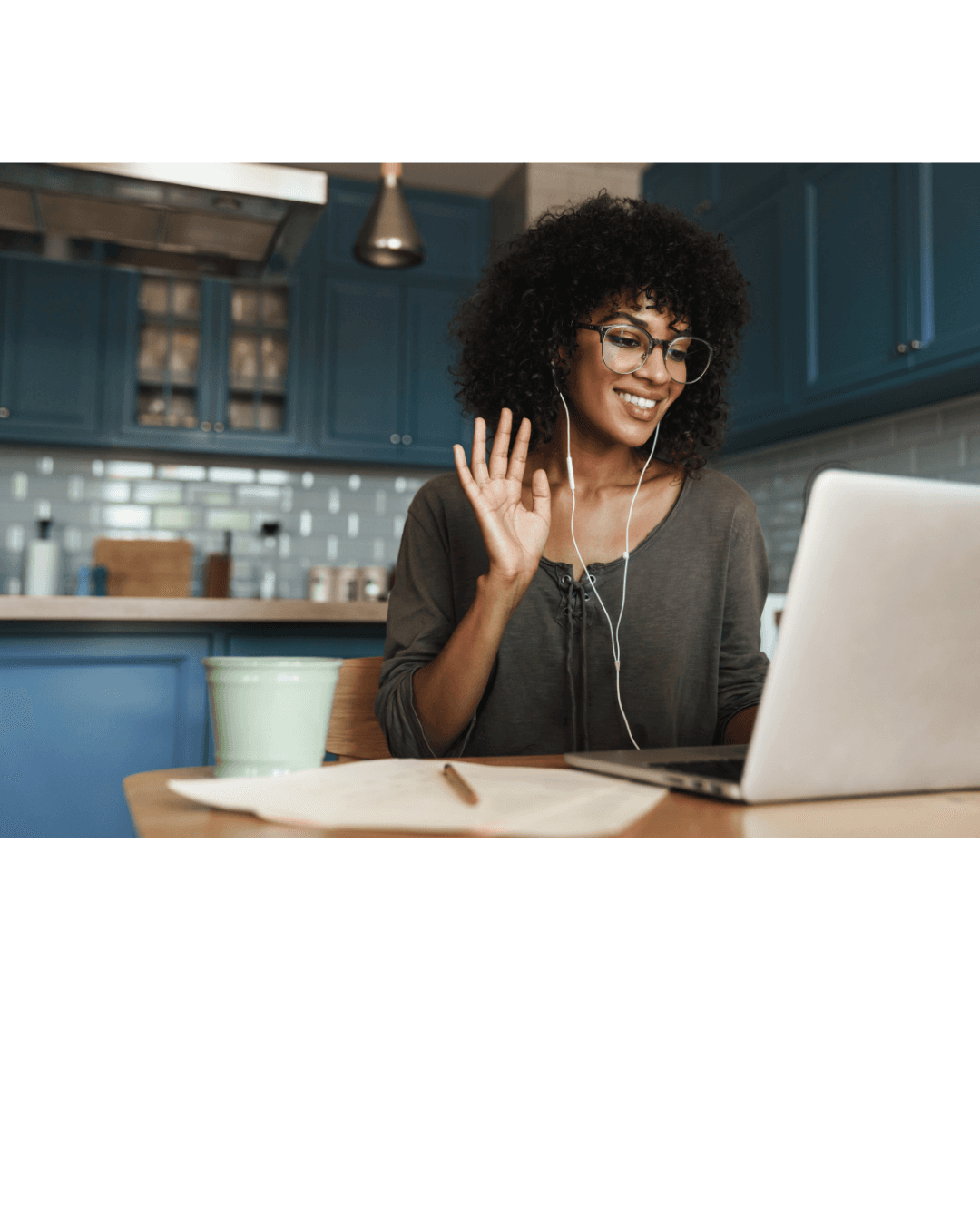 Smiling woman with curly hair and glasses waves at her laptop during a video call.