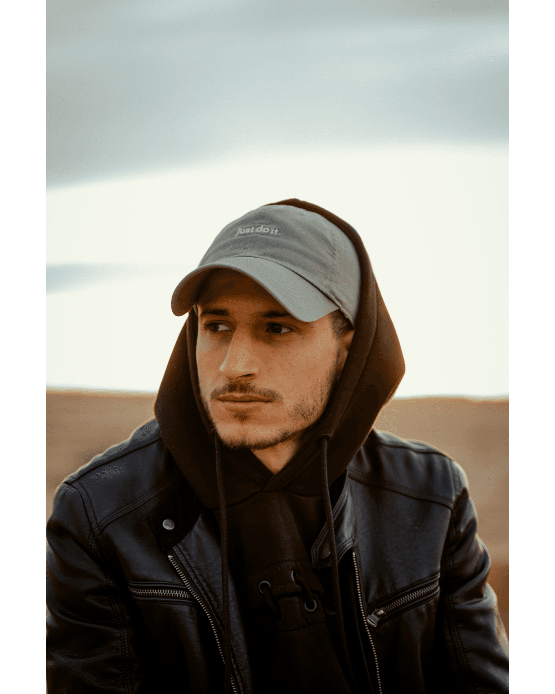 Man in a grey cap and black leather jacket looking thoughtfully aside during golden hour.