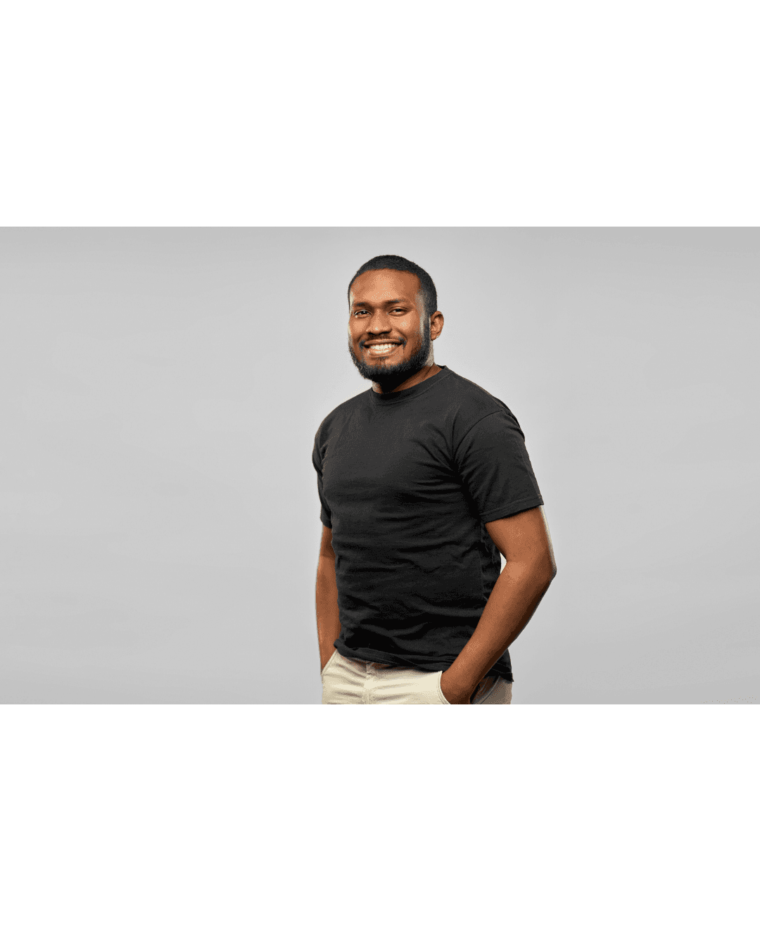 Smiling bearded Black man in a black t-shirt and khaki pants against gray background.